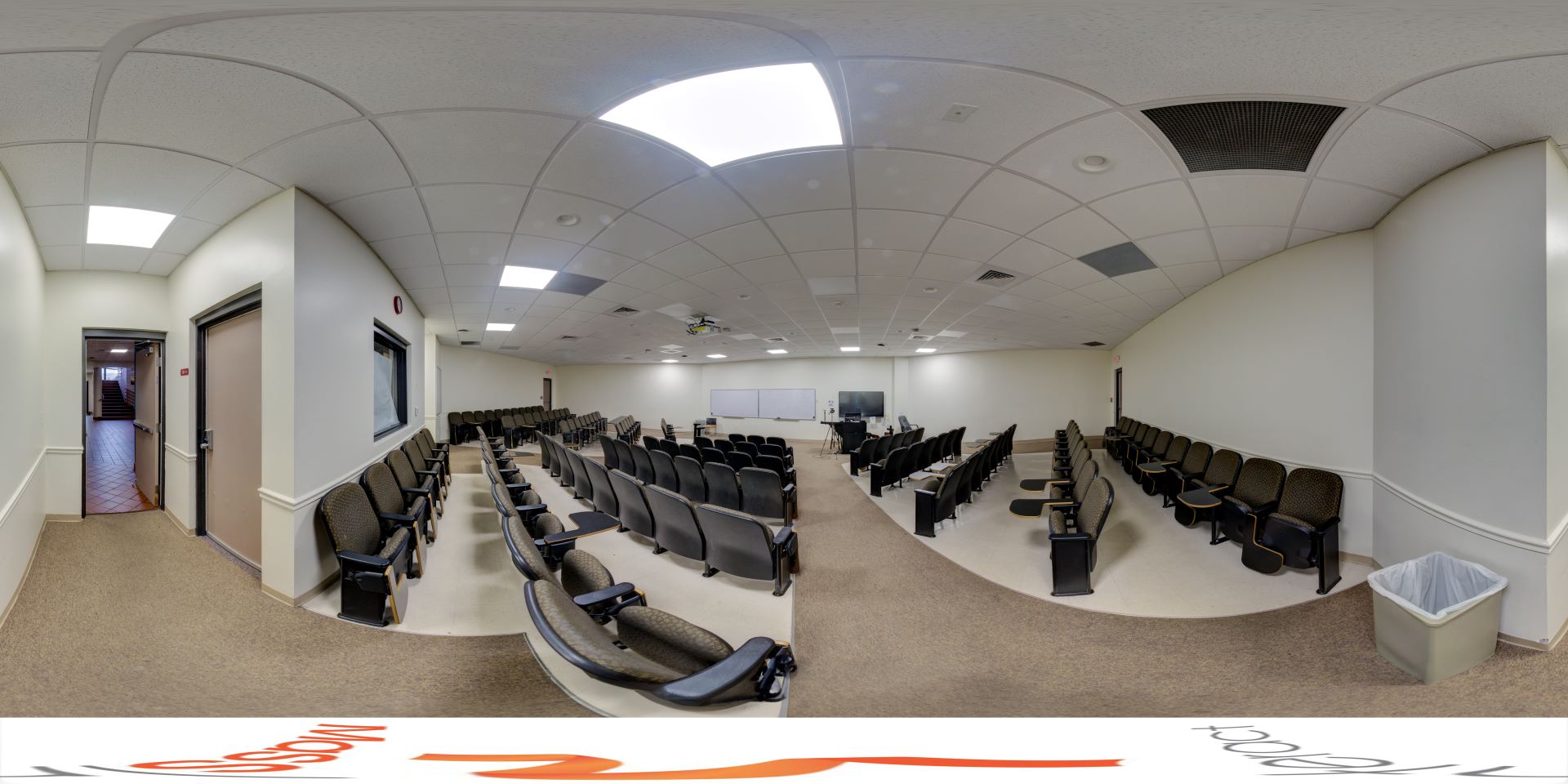 Panoramic view of a large auditorium with rows of black chairs, curved white ceiling, and equipped with multimedia presentation facilities, with an entrance door at the side